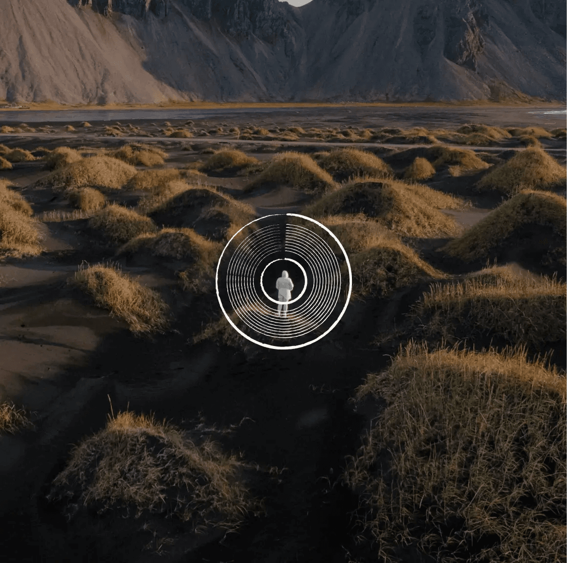 A lone figure among grassy dunes below dark mountains, inside a white concentric-ring emblem