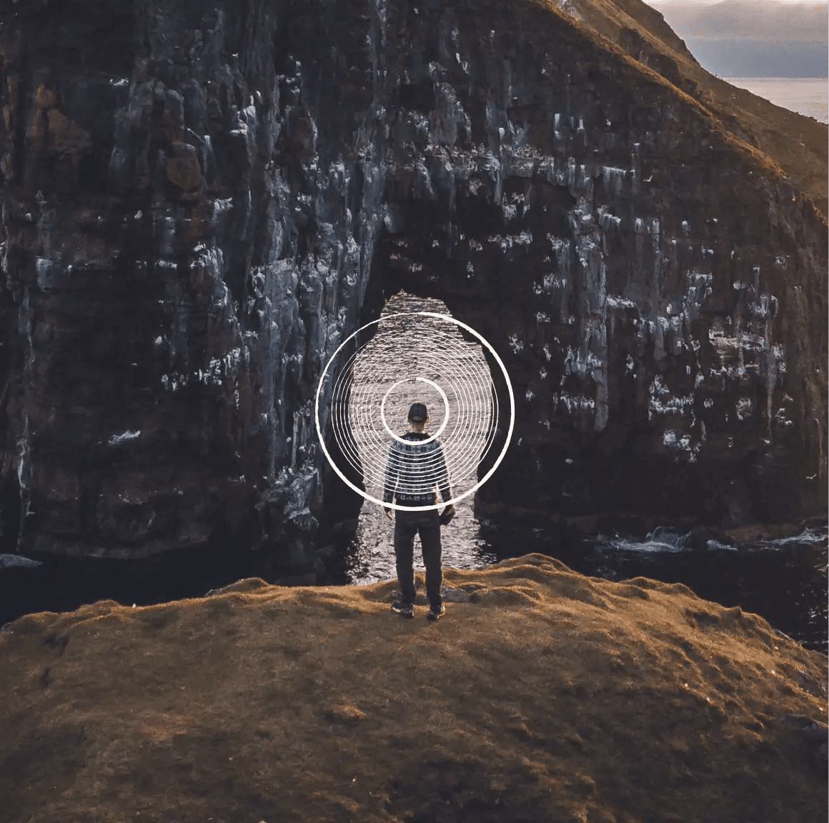 A person standing on a cliff edge facing a sea arch, framed by white concentric rings
